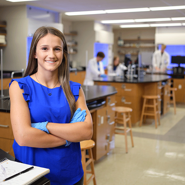 A female student with brown hair and wearing a sleeveless blue dress, her arms crossed and with blue latex gloves on her hands, stands in the foreground with a science laboratory visible behind her. People in white lab coats are visible working in the background.